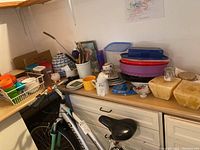 Wide view of countertop and cabinet showing assortment of kitchenware: jars, ceramic containers, plastic bowls, decorative china plates, and kitchen utensils, along with an electric yogurt maker.