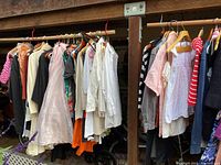 Photo of assorted ladies tops including blouses and shirts hanging on a wooden rod, featuring various colors such as white, pink, beige, and patterns.
