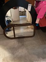 Photo showing two mirrors placed on the floor. The larger round mirror has a black frame and the smaller rectangular mirror has a wood frame.