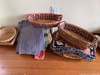 Photo shows a variety of folded linens stacked on a wooden surface next to three oval woven baskets with red, green, and natural wood colors.