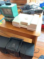 Photo of table with Fisher-Price record player closed, time clock, printer with cords on top, and JVC boom box speakers on floor below.