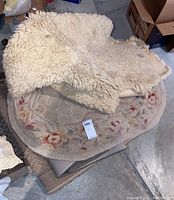 Stacked round beige floral rug and white sheepskin rug, captured on concrete floor with surrounding boxes and items.