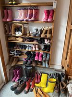 Multiple pairs of children's and adult boots arranged on five wooden shelves. Includes pink, purple, floral rain boots, and brown, black, and gray boots.