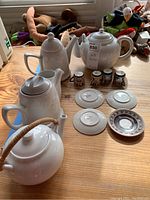 Four white ceramic tea pots of differing shapes arranged on a wood table, alongside four small decorative corked cups and four white ceramic saucers, including one with a decorative dark brown rim.