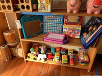 Wide shot of an entire shelf showing various soft toys, educational games, wooden blocks, and musical instruments including drums and a spinning toy.