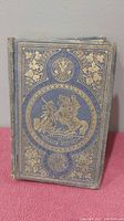 Front cover of a worn hardcover book with blue cloth and gold embossed decorative designs showing a mounted knight and floral elements.