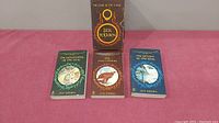 Three softcover books from the Lord of the Rings series by JRR Tolkien, standing on a pink surface with their illustrated covers visible.