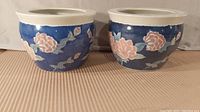 Two blue ceramic flower pots side by side showing full view with floral decorations on a striped surface