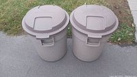 Two beige Rubbermaid Roughneck garbage cans with lids standing on pavement and next to grass.