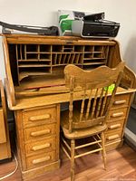 Front view of oak roll-top desk open, showing pigeonholes and writing surface with chair in place.