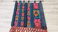 Full view of large tapestry laid flat showing rooster and geometric patterns in multiple colors, hanging tabs at top and fringed bottom edge.