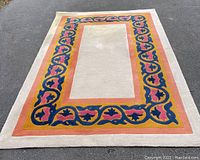 Full top view of the rectangular wool pile rug showing central cream field, orange and gold borders, and floral patterns in navy blue and pink.