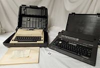 Both electric typewriters displayed open with respective hard cases behind and manuals in front