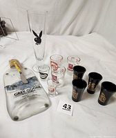 Full view of assorted barware including Grey Goose vodka cheese tray with spreader, various shot glasses including black Wild Spirit and clear ones with red and black logos, and tall Playboy pilsner glass.