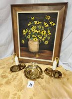 Photo showing painting of black-eyed Susans in ceramic jug framed, pair of brass candle sconces with candles, and gold leaf damask tablecloth beneath them
