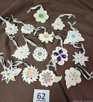 Photo showing all 14 handmade soft polymer flower and star shaped Christmas ornaments arranged on brown fabric. Each ornament is lightweight, white with colorful gem and mirror decoration, featuring silver hanging ribbons.