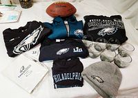 Photo showing all apparel items including quarter-zip pullover, hoodie, short sleeve shirts, child's long sleeve tee, knit hat, along with Wilson football, seven glasses, and Eagles Care Summit journal and pen.