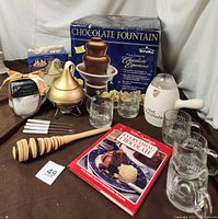 Chocolate fountain in box along with Hershey's Kisses fondue pot, clear mugs, hot chocolate pitcher, wooden whisk, cookbook, and Godiva chocolate box arranged on table.