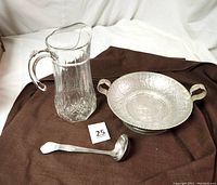 View of the pitcher, hammered silver bowl, and metal ladle together on a brown cloth background to show the full lot.