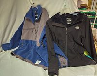 Two North Face jackets displayed on hangers on a white backdrop. The women's black jacket with velour lining and hood is on the right, the men's blue and grey fleece and nylon jacket is on the left.