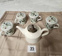Photo showing four floral print mugs each with a ceramic lid, and a white ceramic teapot with a metallic lid placed on a beige checked cloth.