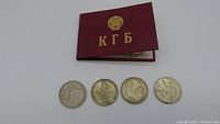 Four Soviet 1 Ruble commemorative coins aligned with KGB identity card booklet with gold lettering and emblem on cover.