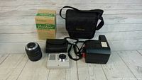 Lot for sale showing Canon digital camera with carrying case, Nikon lens, Polaroid instant camera and Fujifilm Printpix paper box on flooring.