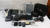 Photo showing a variety of phones, a digital camera, video camera, and bags including a projector in the background.