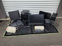 Lot of Dell Optiplex desktop PCs, Dell monitors, UPS units, keyboards, speakers, and mouse laid out on black mat outside storage unit.