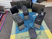 Three Dell Optiplex desktop computers on a colorful cloth with power cables and six assorted monitors in the background, plus one APC Back-UPS device in front.