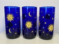 Front view of three cobalt blue glass tumblers showing sun, moon, and stars designs in gold and blue.