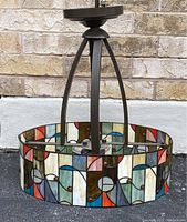 Pendant light with stained glass shade showing geometric abstract design, colors include blue, green, amber, white, and red. Metal frame dark colored with tripod supports.