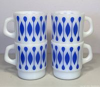 Four stacked vintage white milk glass mugs with vertical blue teardrop patterns and square handles.