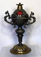 Front view of antique ornate metal oil lamp showing detailed metalwork and red glass inserts.