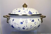 Side angle of vintage blue and white floral enamel casserole pot showing copper handles and lid with brass finial.