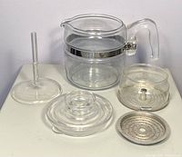 All individual parts of the coffee percolator shown disassembled on table including glass pot, lid, stem, metal filter plate and basket