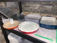 Photo showing four glass pie dishes, white Corning-French rectangular dish, cast iron baking dish, Le Creuset and Sara Dolce ceramic baking dishes on shelf against stone wall