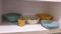 Five bowls on white shelf: turquoise scalloped bowl, yellow flower-patterned small cup, white scalloped bowl, large yellow fluted bowl, turquoise rectangular scalloped dish.