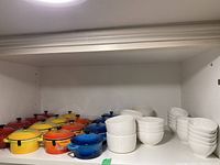 Overall view of 16 miniature colorful cooking pots and 24 small white ceramic bowls stacked beside them on a white shelf.