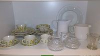 Full view of a shelf containing floral patterned cups, saucers, bowls, a white ceramic jug, clear textured glass candy dishes and plate with bug/bee motifs.