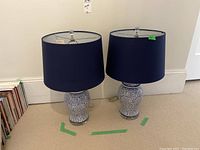 Pair of ceramic table lamps with white and blue swirling pattern bases and navy blue drum lampshades, both approximately 25 inches tall, placed on carpeted floor near wall.