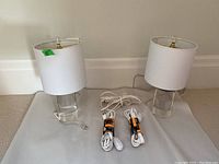 Two glass base lamps with white fabric drum shades approximately 15 inches tall with three white Noma extension cords coiled nearby. Lamps have clear cords and brass hardware at tops.