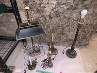 Five vintage lamps arranged on basement floor near metal shelving, four have metal bases, one wooden base. Three lamps missing shades, some wear and damage noted.