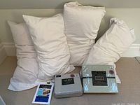 Four white queen pillows with covers, two unopened Ralph Lauren sheet sets, and unopened pillow protectors package