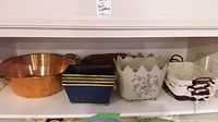 Shelf displaying the copper basin, blue rectangular planters, white floral scalloped planter, and white baskets with brown handles.