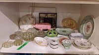 Wide view showing plates, glass flower frogs, pink pig figurine, framed item, and other ceramic dishes on a shelf