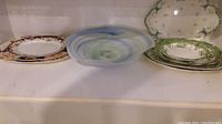 Three types of platters and a glass bowl arranged on a shelf. Platters have floral and green rimmed designs. Bowl is blue-green swirl glass.
