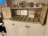 Shelf display showing two large framed prints, a framed oil painting, a wooden 'SHOP like you mean it' sign, and eleven small framed entomology prints on display. Plates on shelf above