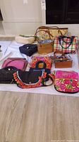 Full lot photo showing variety of 12 purses and makeup bags arranged on floor including wicker, fabric, and woven styles. Various colors including pinks, black, multicolor and natural wicker tones.