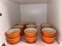 Sixteen ceramic ramekins arranged in groups on a shelf, showing matching orange gradient colors and ribbed sides.